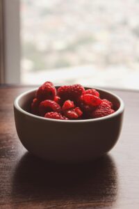 Close-up of vibrant red raspberries in a bowl on a wooden table by a window.